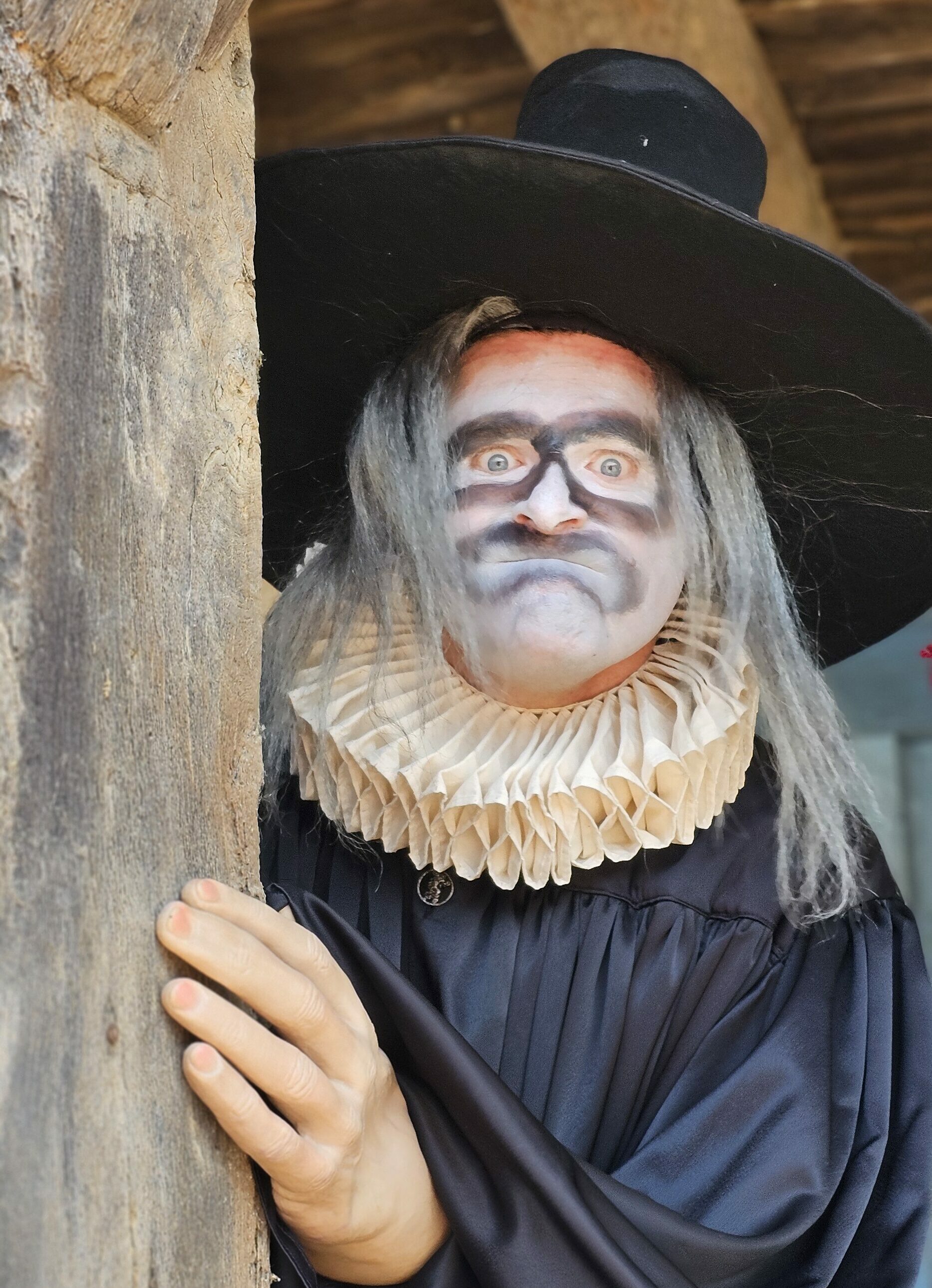 Portrait de près d'un comédien costumés avec une robe noire, un chapeau noir, une perruque grise et un maquillage noir et blanc.