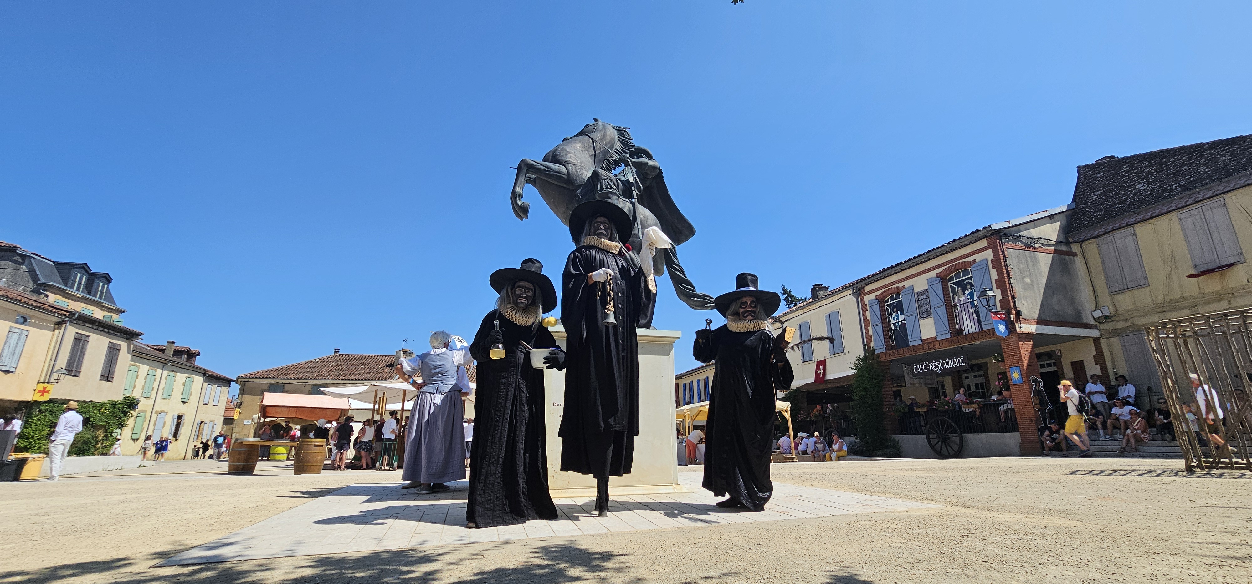 Trois comédiens costumés avec de longues robes noires et des chapeaux noirs au centre d'une place.