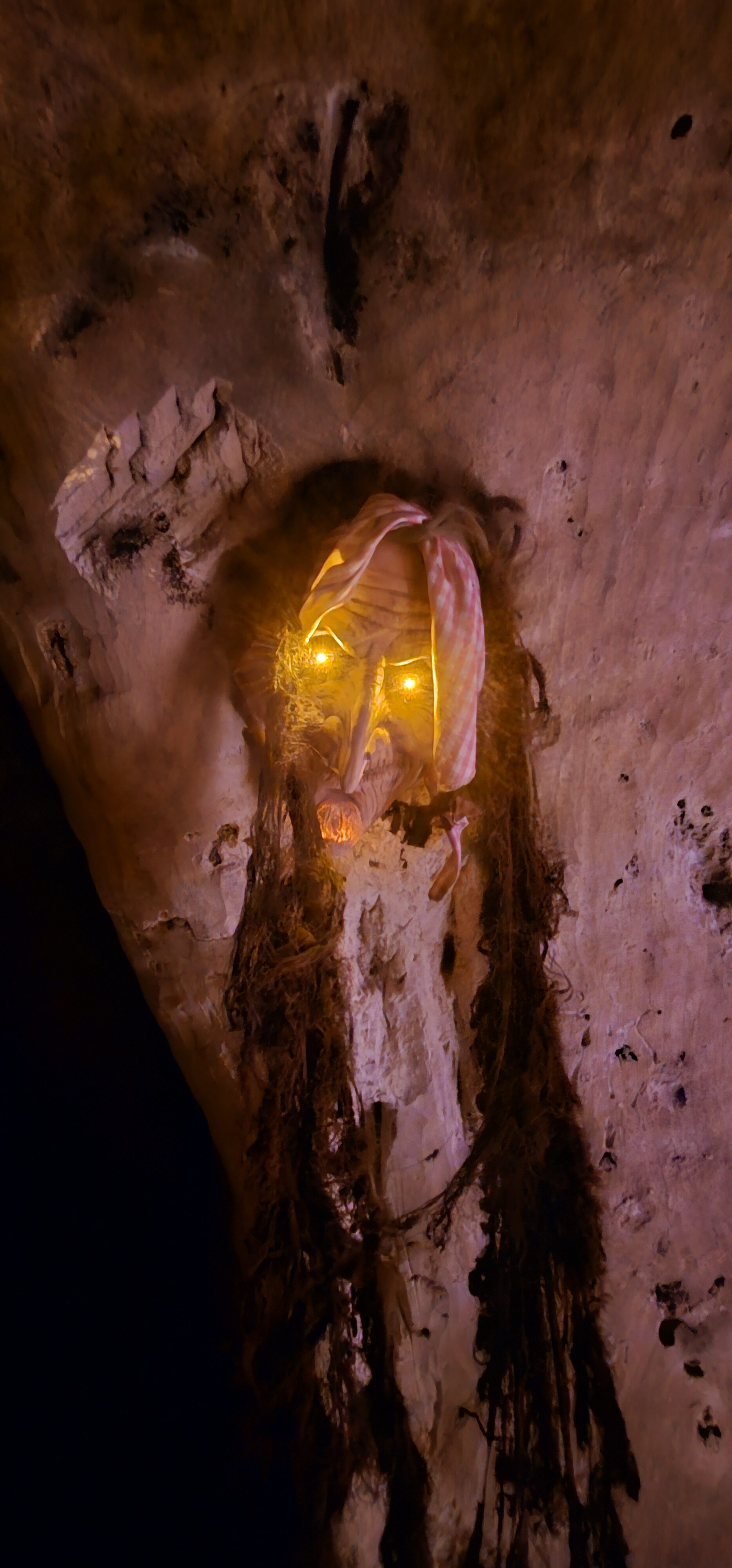 Masque effrayant accroché sur un mur en pierre avec des yeux lumineux.