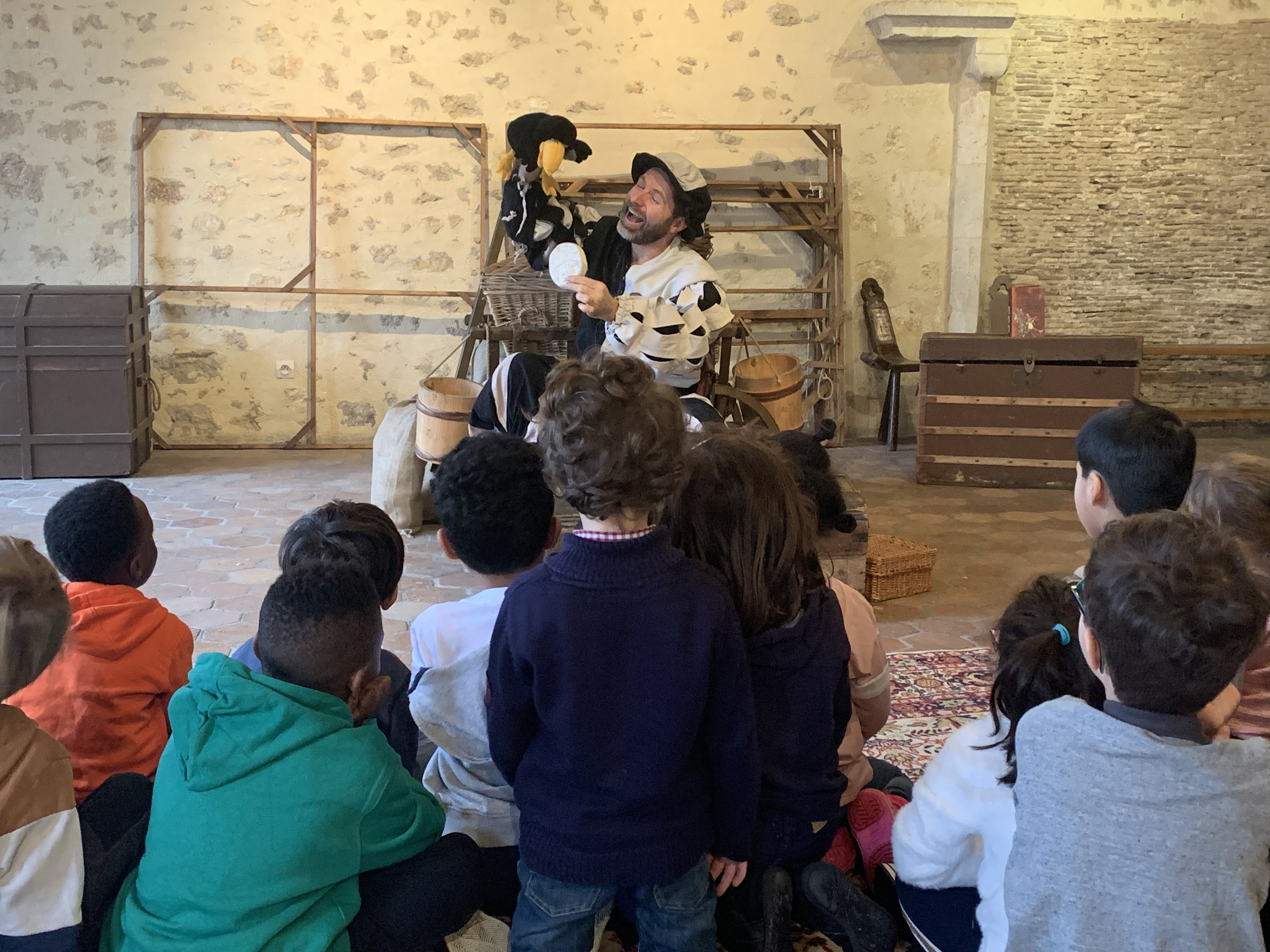 Groupe d'enfants de dos admirant un comédien déguisé et manipulant une marionette.